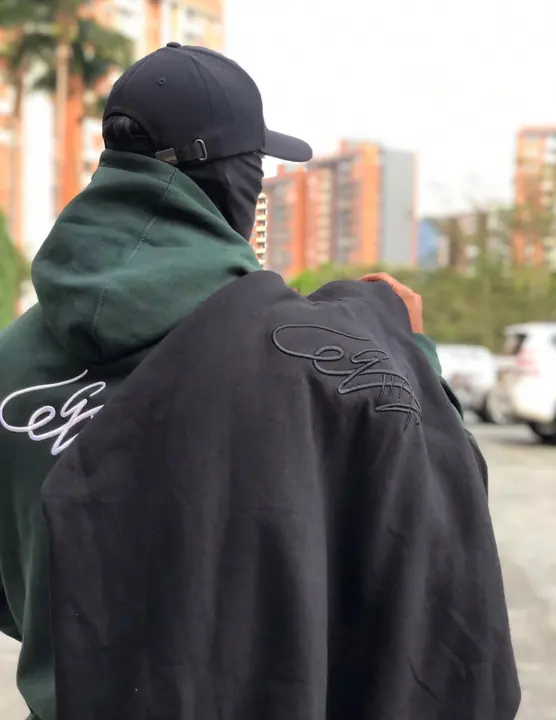 Hoodie verde puesto y hoodie negro tonal al hombro, en la calle